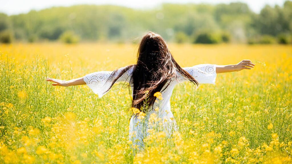 A woman with long hair wearing a white dress with lace sleeves stands in a field of yellow flowers, spreading her arms wide, embodying web design psychology principles of harmony and balance.